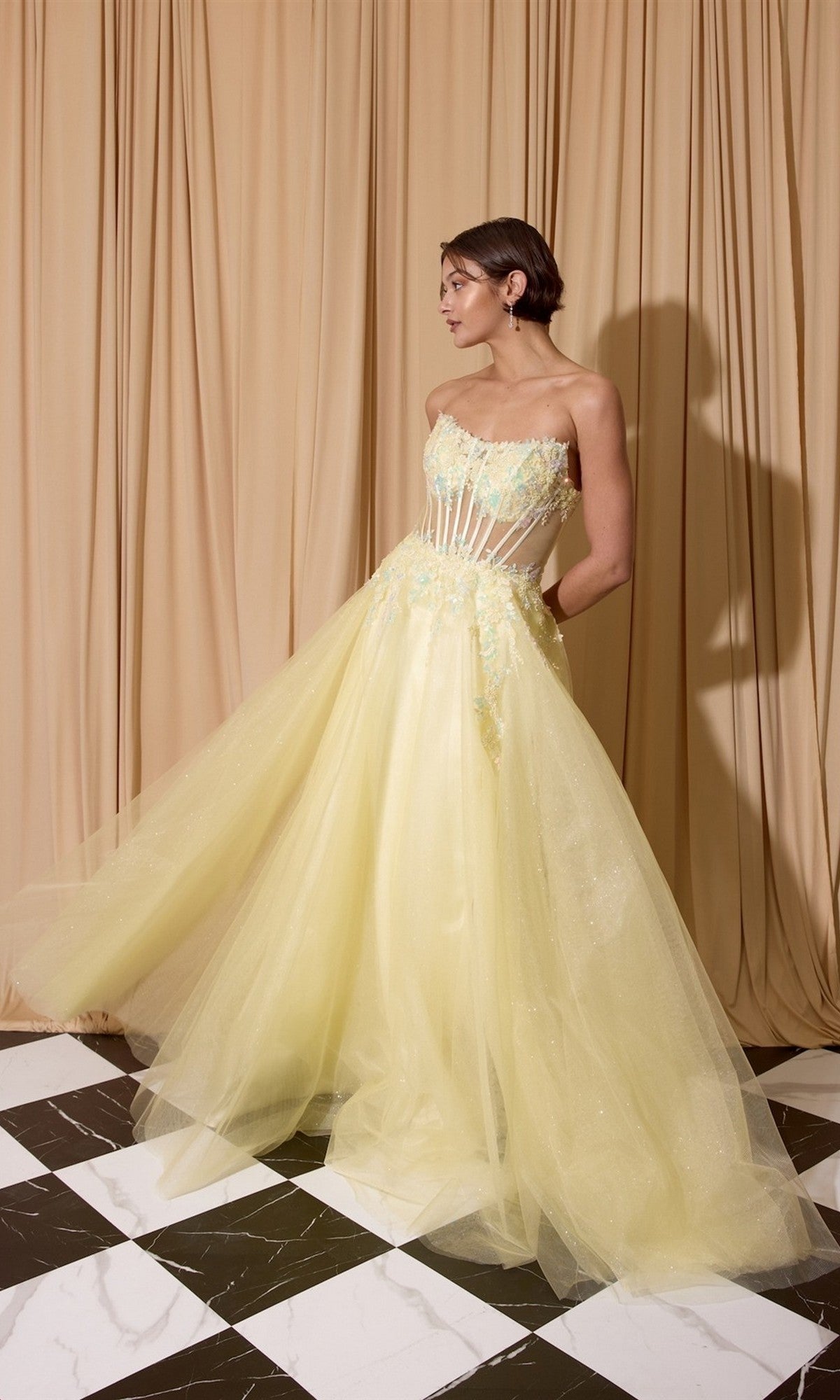 A woman stands on a black and white checkered floor, wearing the Eureka EK208 long prom dress in pastel yellow glitter tulle with a strapless semi-sheer corset bodice and beaded floral appliqué, in front of beige curtains.
